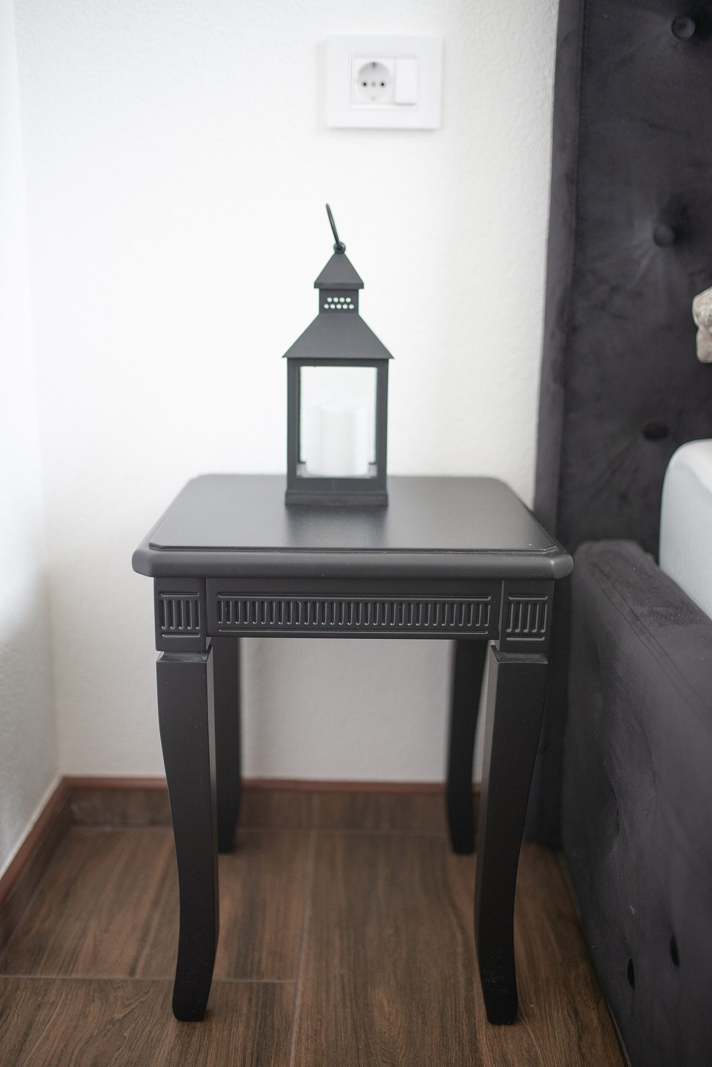 A black side table beside a bed against a white wall, with a socket on the wall above it