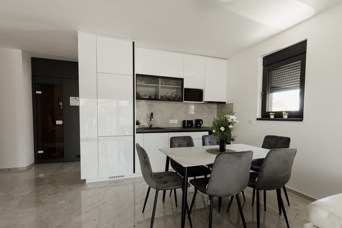 A bright kitchen diner with a white stone table and six grey chairs set close to the kitchen run
