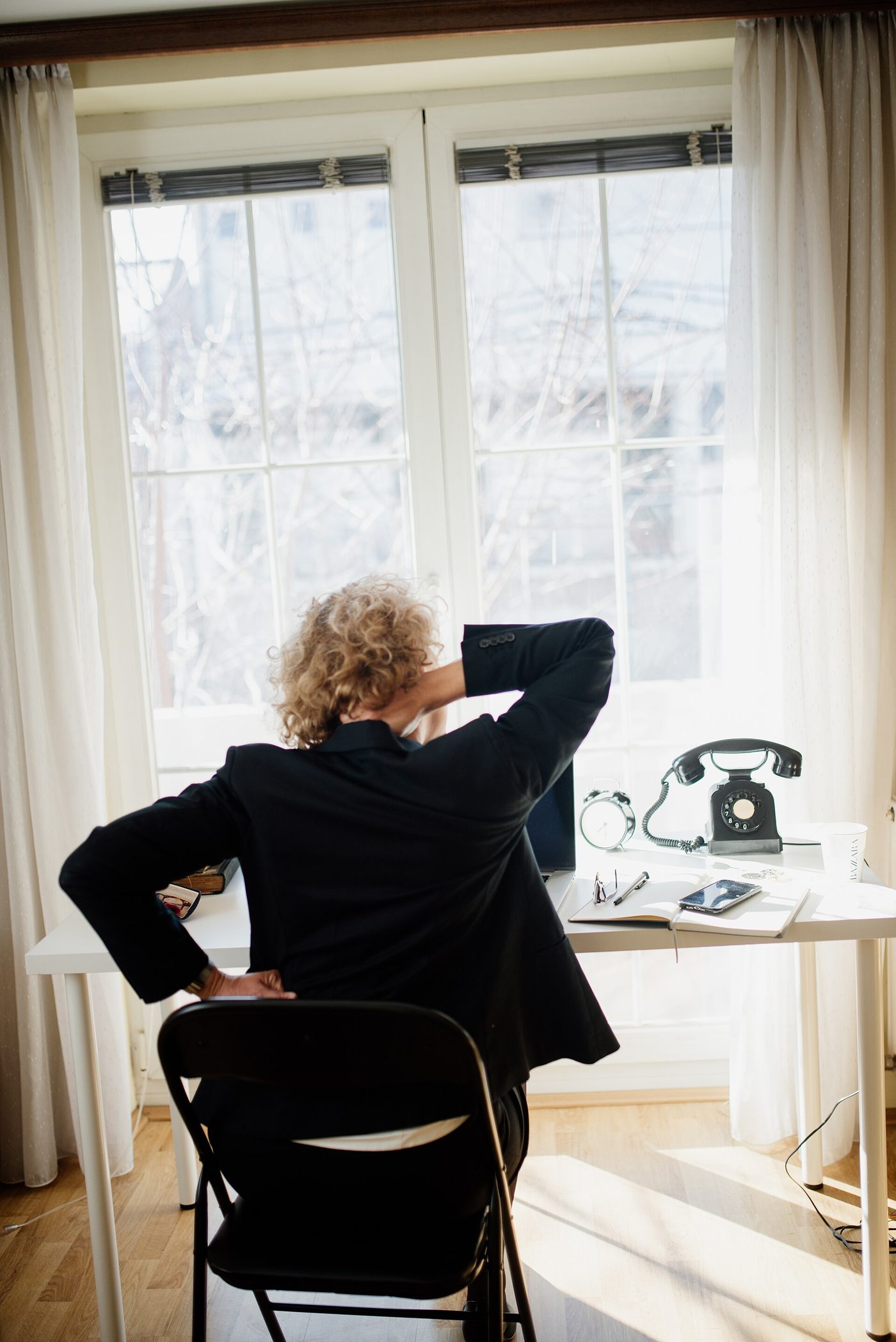 A person sitting at a white desk placed directly in front of a bright window, seen from behind