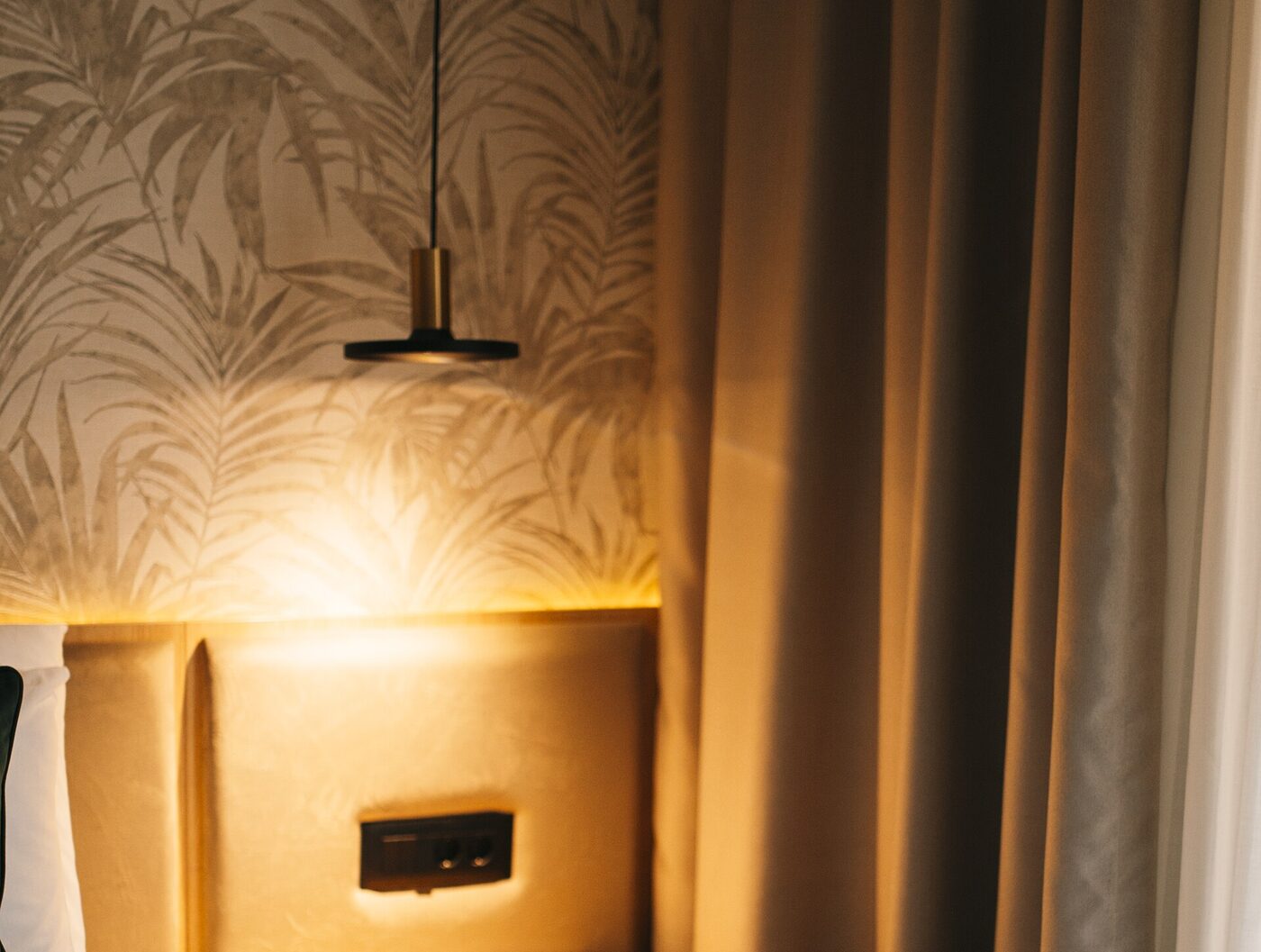 A warm pendant lamp washing light down a patterned wall above a bed, with a socket set into the headboard