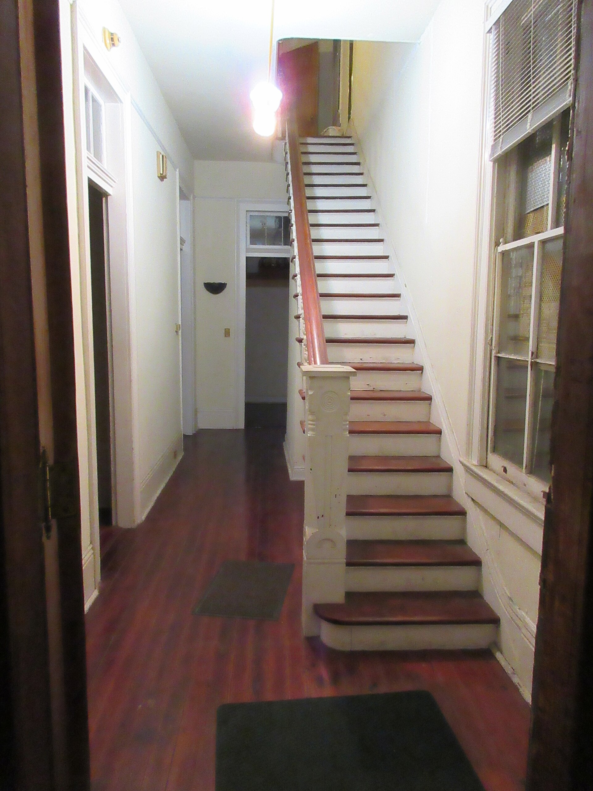 A dim, dated house hallway with a staircase and bare walls
