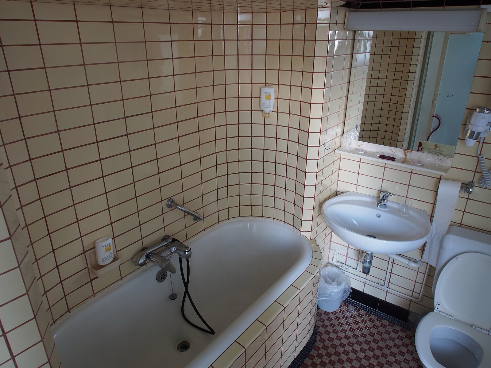 The original bathroom: cream square tiles with red grout, a tub in a curved tiled alcove, a wall hung basin with exposed pipes, a mirror with a light bar and a toilet