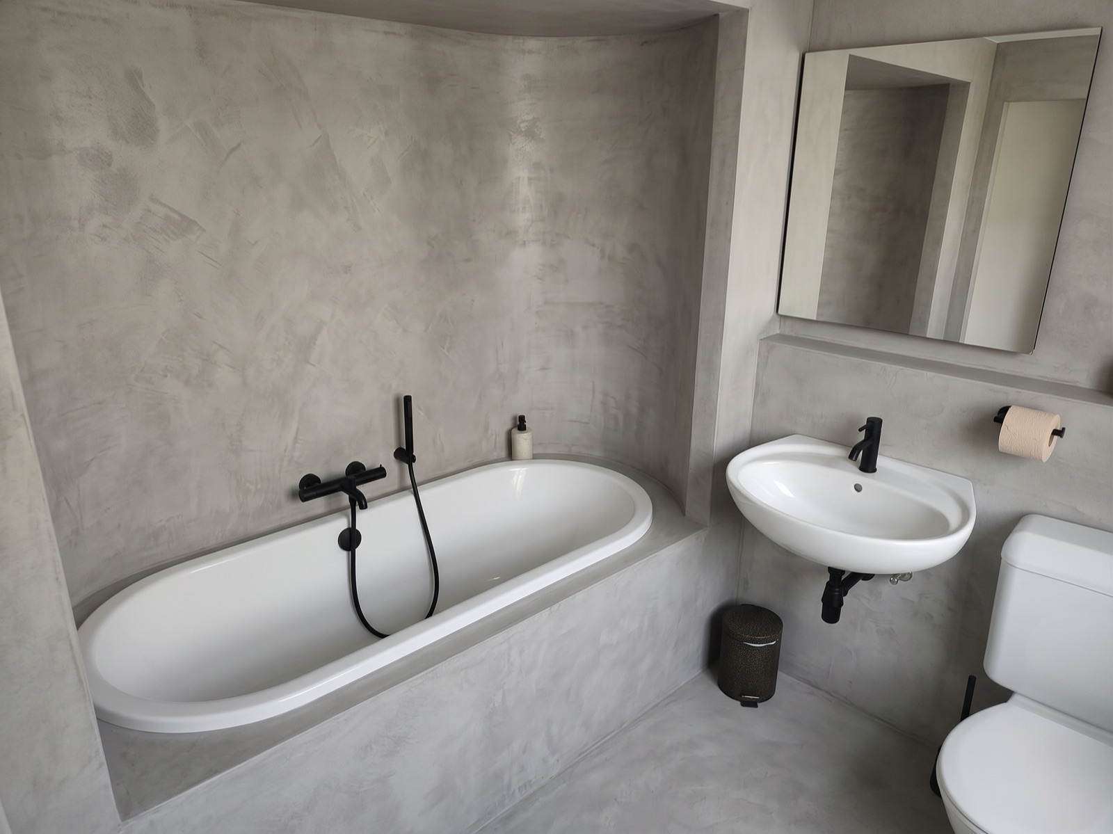 The same bathroom redesigned in grey microcement, with black taps, the same tub position, basin, mirror and toilet