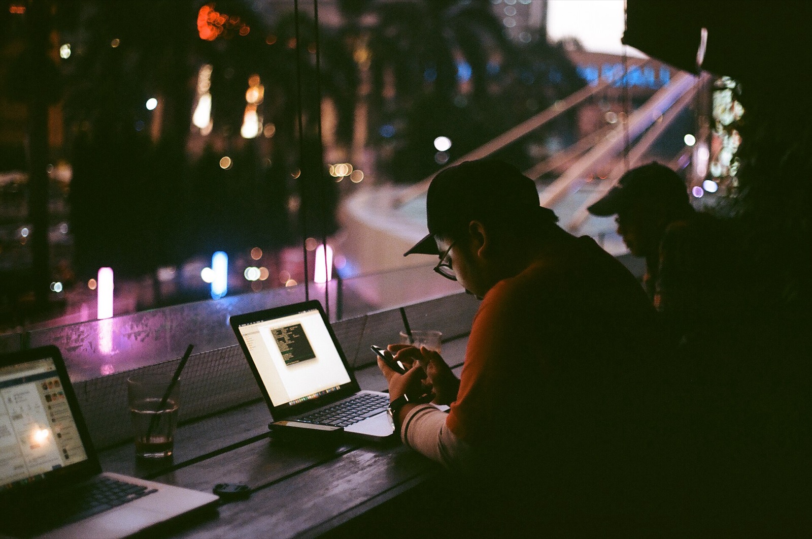 A person in a cap using a phone at a laptop desk at night with city lights behind