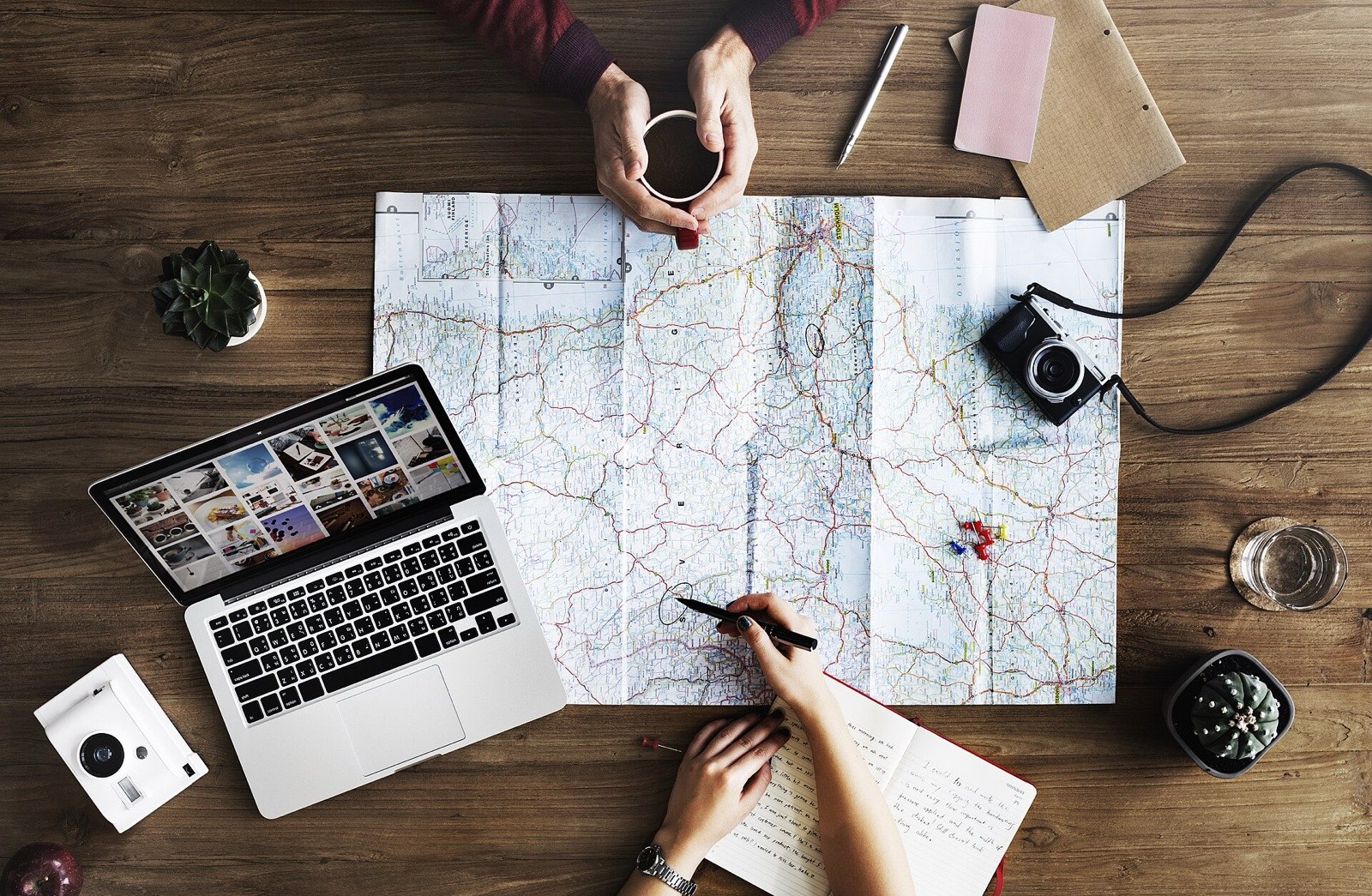 Two people planning a trip over coffee with a paper map, a laptop and a camera on a wooden table