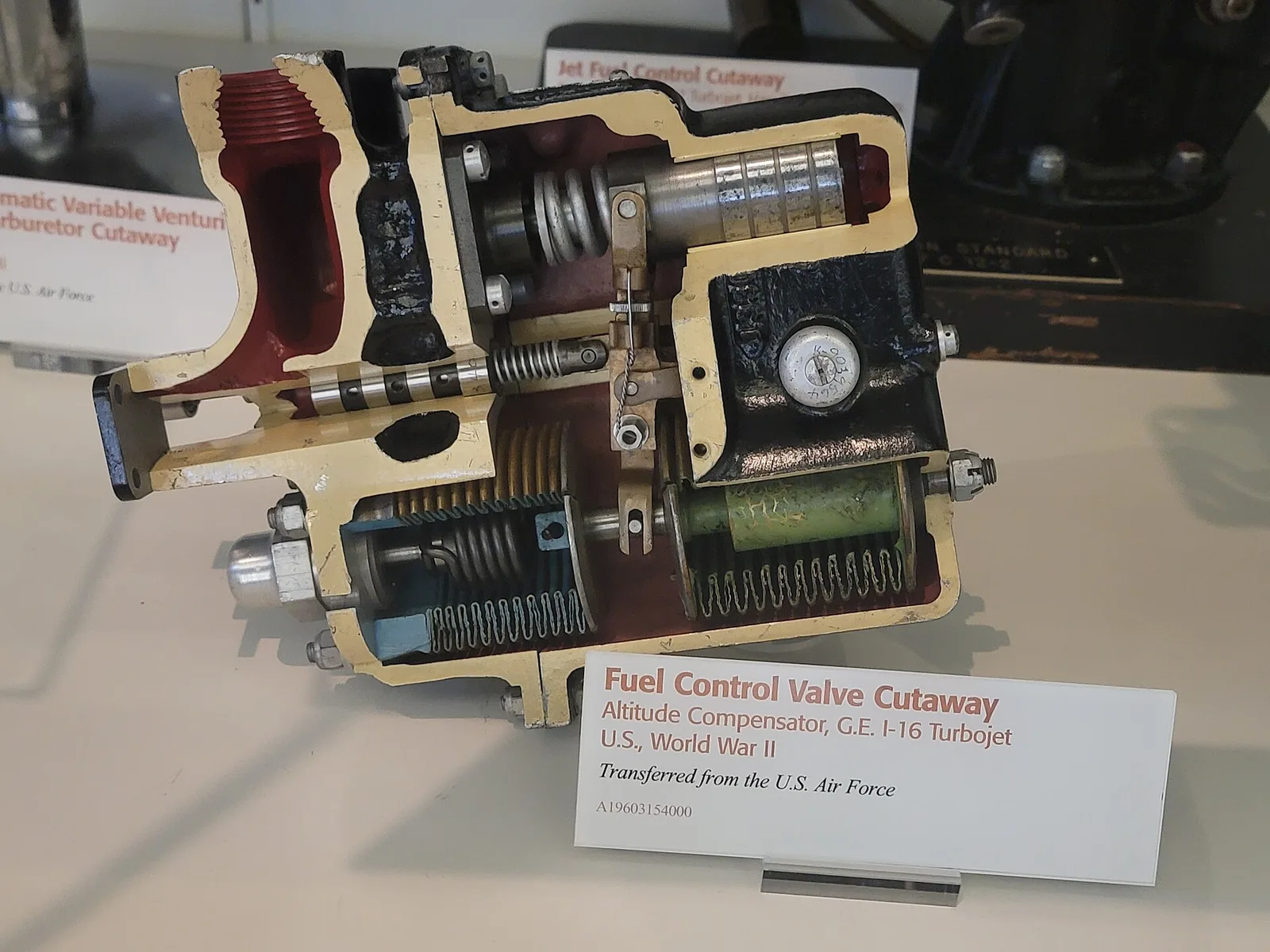A 1940s jet engine fuel control valve shown as a museum cutaway, with the cut faces painted so the internal springs, valves and passages are visible
