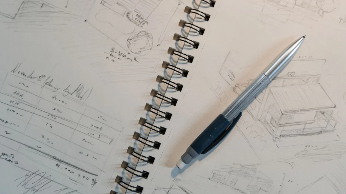 Hand-drawn design sketches and notes filling a spiral notepad, with a drafting pen resting on the page